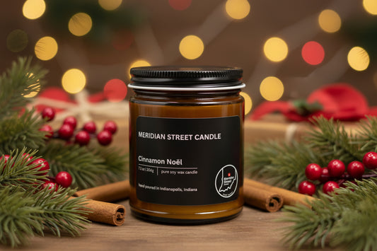 Meridian Street Candle Cinnamon Noel candle surrounded by Christmas decorations on a wooden surface with blurred lights in the background.