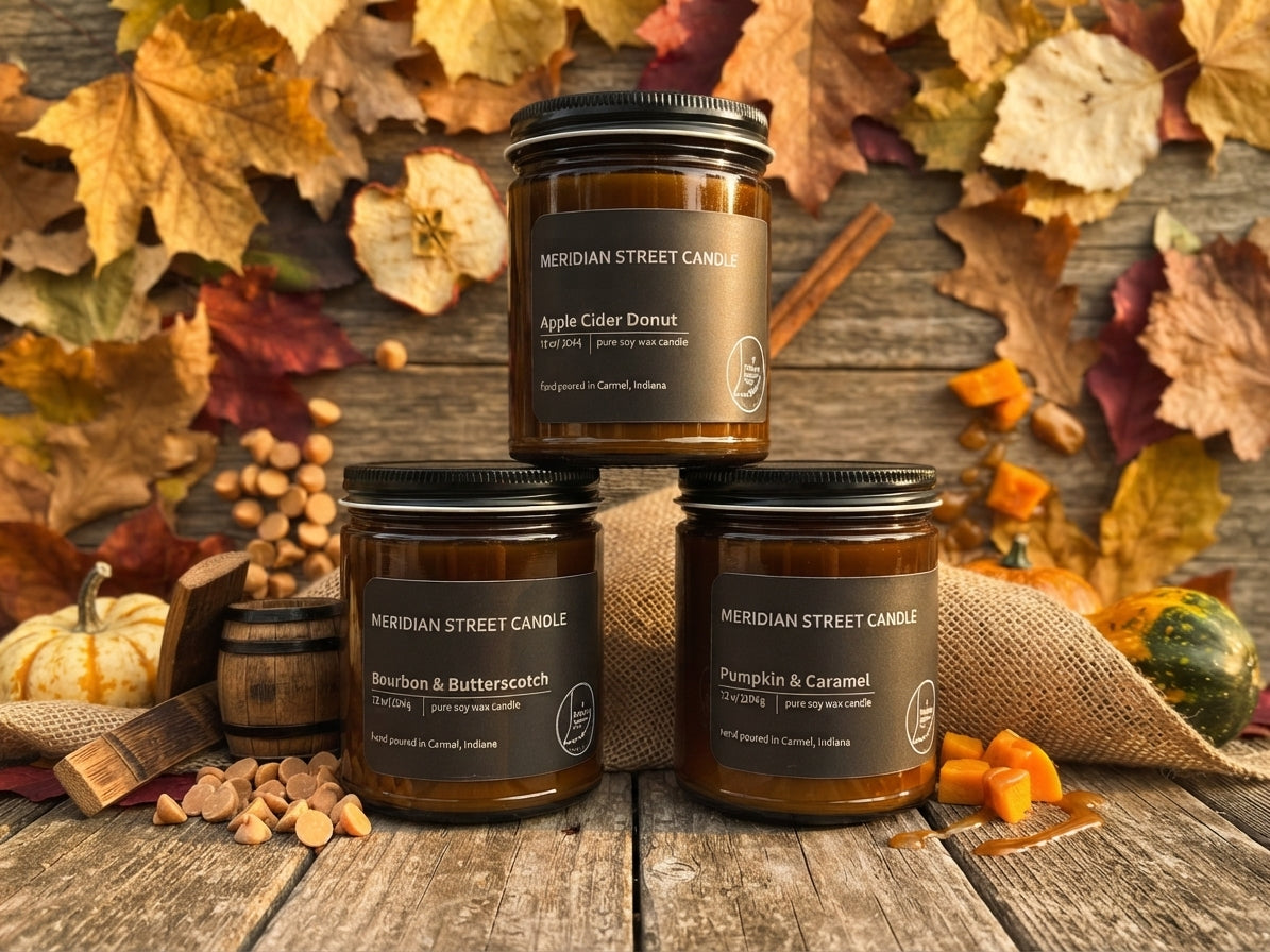 Three amber glass jar candles from Meridian Street Candle are arranged on a rustic wooden surface with autumn leaves, pumpkins, burlap, and cinnamon sticks. The labels show different scents: Apple Cider Donut, Bourbon & Butterscotch, and Pumpkin & Caramel.